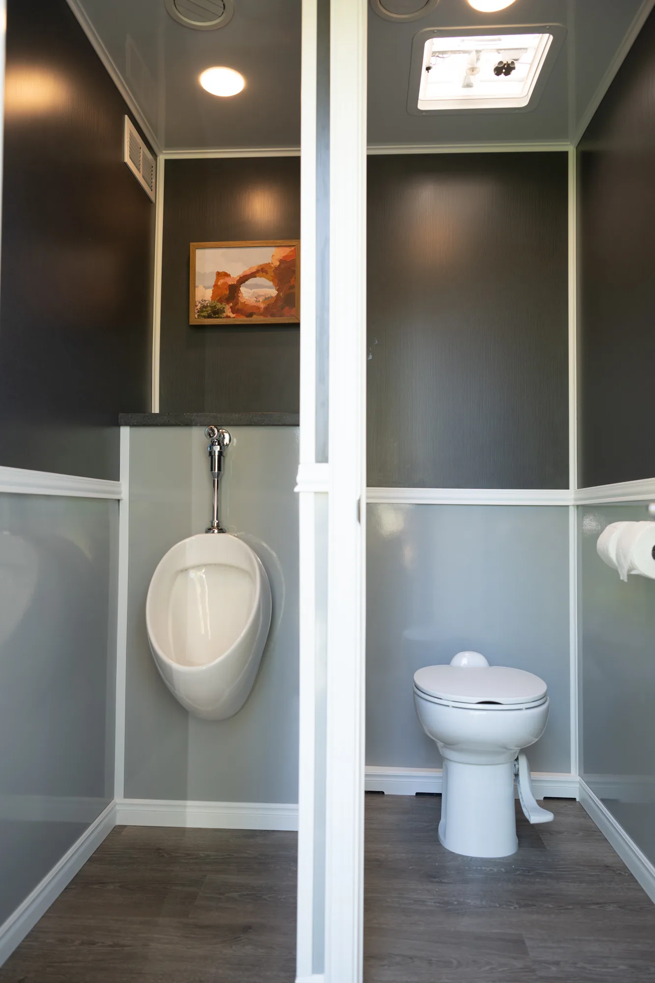 Private stalls inside the luxury restroom trailer with flushing toilet, urinal, and wood-grain floors